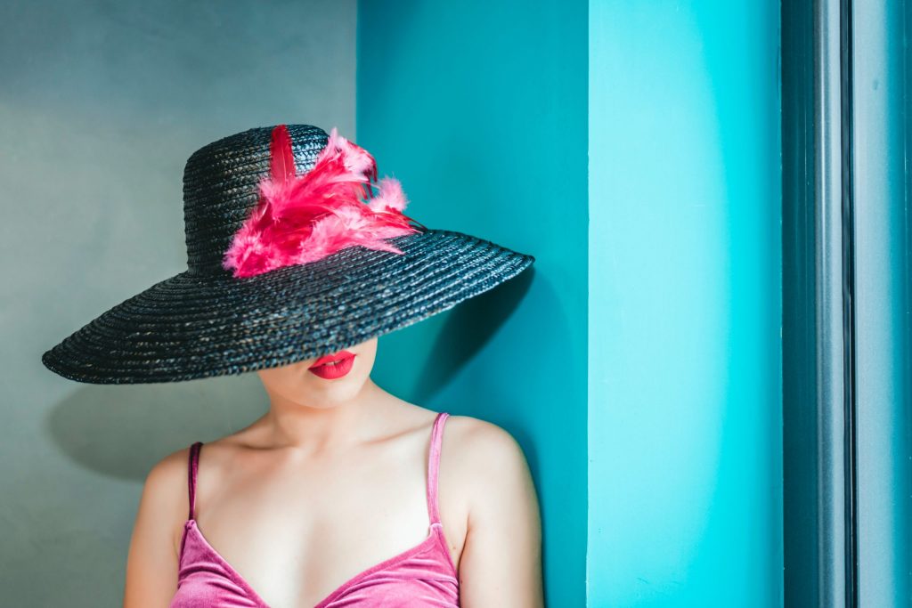 A woman standing in the corner of a room, concealing her hair and face under a fashionable, wide-brimmed hat.