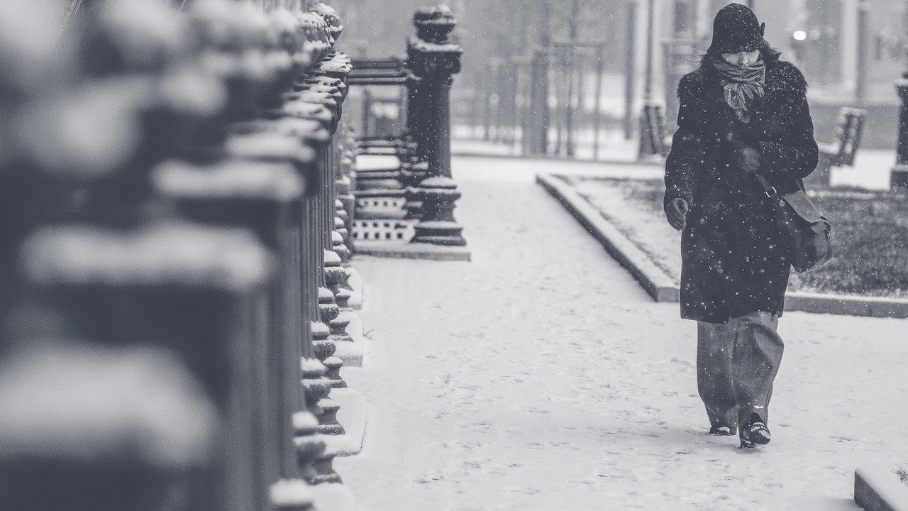 A black and white photo of a woman walking on an urban sidewalk in the snow.